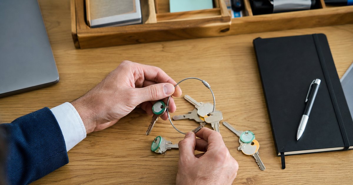 Overhead view of a wooden desk with several small brass keys of different shapes being gathered into a single metal key ring, a notebook and pen beside them