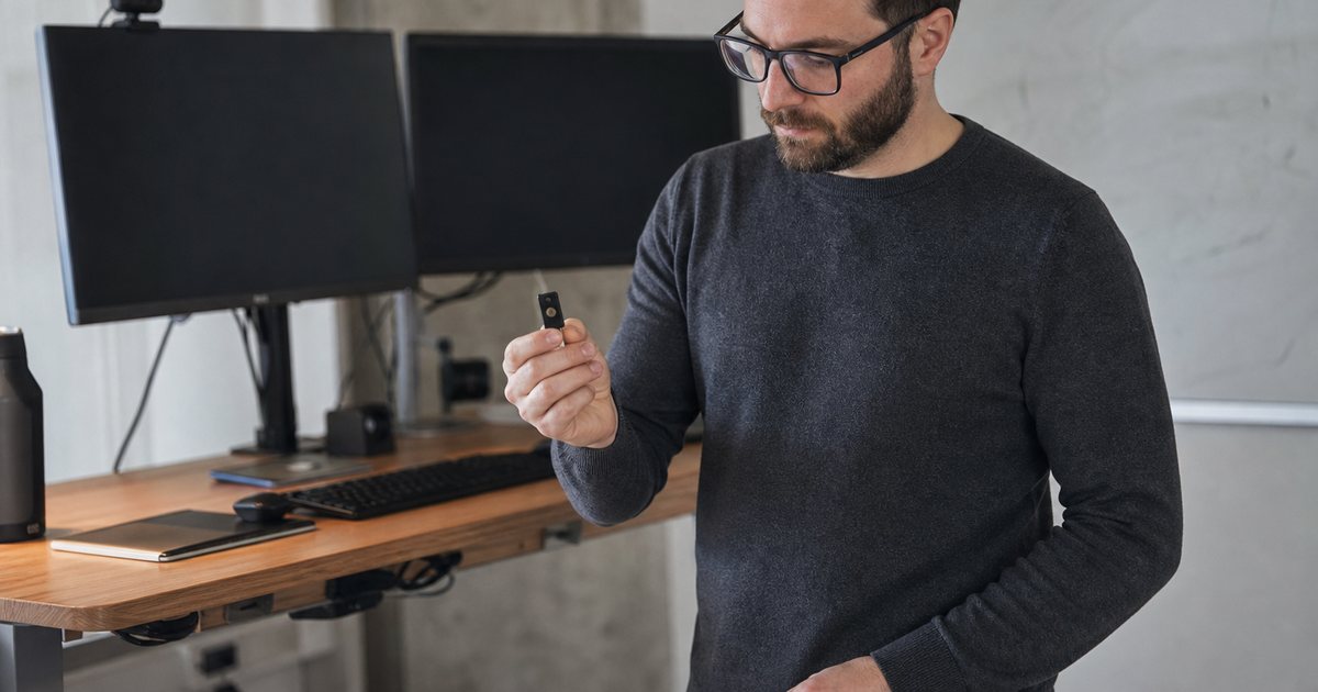 A developer at a standing desk in a small engineering office holding a hardware security key, two monitors switched off behind, whiteboard with erased marks on the wall