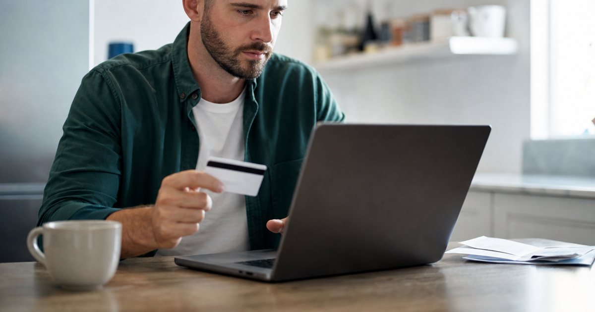 A person at a kitchen table holding a credit card next to a laptop, a cup of coffee and a closed leather wallet, natural window light